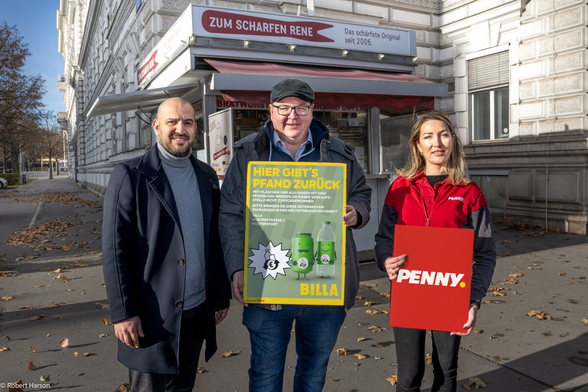 Drei Personen stehen auf einem Bürgersteig vor einem Gebäude. Der Mann in der Mitte hält ein Schild mit dem Text "Hier gibt's Pfand zurück". Die Frau rechts hält ein rotes Schild mit der Aufschrift "PENNY". Alle drei tragen warme Kleidung.