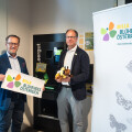 Zwei Männer stehen lächelnd nebeneinander in einem Innenraum. Sie halten ein Schild mit dem Logo von "Blühendes Österreich" und einen Plüschbienen. Neben ihnen steht ein Roll-up-Banner mit dem gleichen Logo und Schmetterlingsabbildungen. Im Hintergrund ist ein Pfandflaschen-Rückgabeautomat zu sehen.