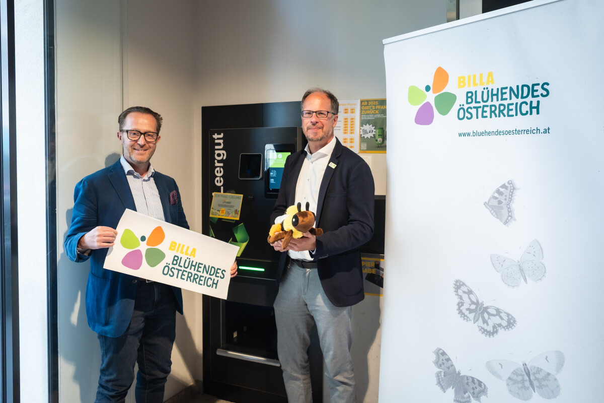 Zwei Männer stehen lächelnd nebeneinander in einem Innenraum. Sie halten ein Schild mit dem Logo von "Blühendes Österreich" und einen Plüschbienen. Neben ihnen steht ein Roll-up-Banner mit dem gleichen Logo und Schmetterlingsabbildungen. Im Hintergrund ist ein Pfandflaschen-Rückgabeautomat zu sehen.