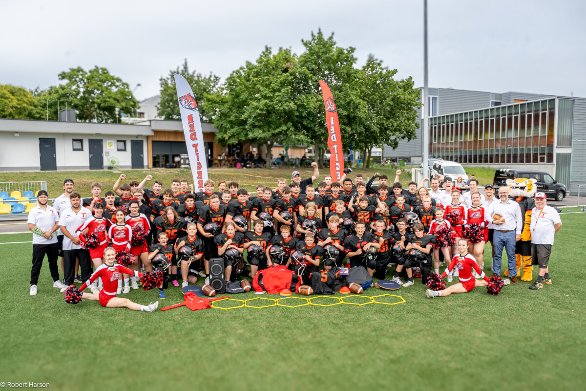 Eine große Gruppe junger American Football-Spieler in schwarzen Trikots posiert auf einem Sportfeld. Cheerleader in rot-weißen Uniformen knien vorne. Im Hintergrund sind Gebäude und Bäume zu sehen. Zwei rote Fahnen sind aufgestellt.