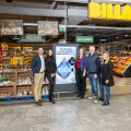 Mehrere Personen stehen in einem Supermarkt neben einem Werbeplakat. Im Hintergrund sind Regale mit Obst und Gemüse sowie Weihnachtsdekorationen zu sehen.