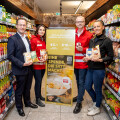 Vier Personen stehen in einem Supermarktgang vor einem Spendenplakat, umgeben von Regalen mit Getränken. Drei tragen rote Jacken mit einem Logo, die andere Person trägt einen dunklen Anzug. Sie halten Lebensmittelpackungen in den Händen und lächeln in die Kamera.