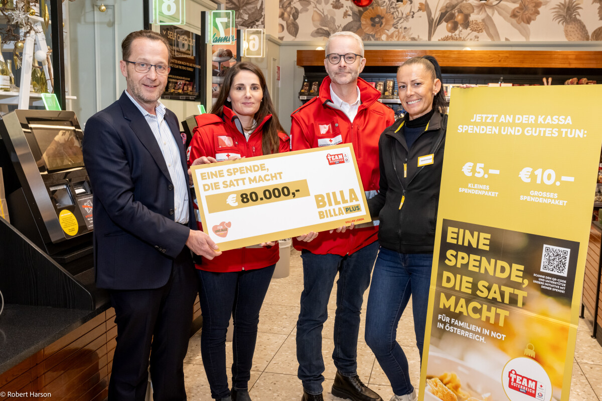 Vier Personen stehen in einem Geschäft und halten Schilder mit der Aufschrift „Eine Spende, die satt macht“ und einem Betrag von 80.000 Euro. Sie tragen Jacken mit Logos und lächeln. Im Hintergrund sind Kassenbereiche zu sehen.