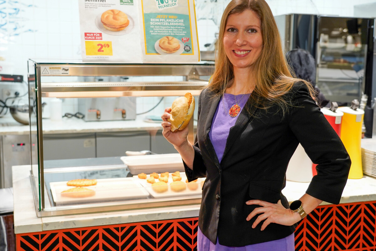 Eine Frau in einem lila Kleid und schwarzen Blazer hält ein Sandwich in einem Imbiss. Im Hintergrund sind weitere Sandwiches unter einem Schild mit der Aufschrift "Vegavita Schnitzelsemmel" zu sehen. Auf einem weiteren Schild steht "100% pflanzlich".