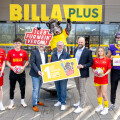 Eine Gruppe von sechs Personen steht vor einem BILLA PLUS-Geschäft. Zwei Personen in der Mitte halten ein Schild mit der Aufschrift "Lebe! für mein Verein" und "BILLA PLUS". Rechts steht ein Maskottchen in einem Kuhkostüm. Drei Personen tragen Sportbekleidung.