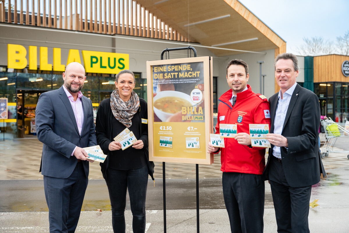 Vier Personen stehen vor einem Supermarkt, halten Flyer in der Hand und lächeln. Ein Plakat in der Mitte wirbt für eine Spendenaktion. Im Hintergrund ist das Logo von "Billa Plus" zu sehen.