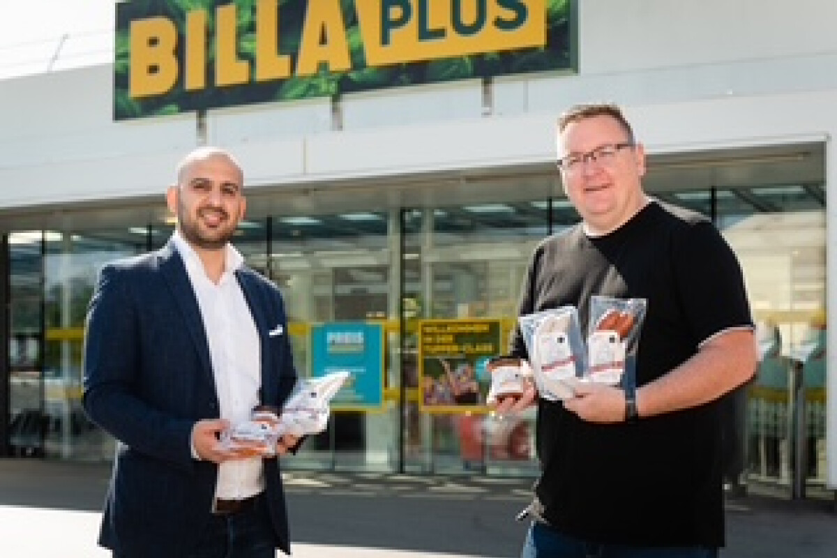 Zwei Männer stehen vor einem Billa Plus-Geschäft und halten Produkte in den Händen.