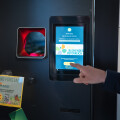 Eine Person bedient einen Leergutautomaten in einem Geschäft. Auf dem Bildschirm wird eine Kampagne für „Blühendes Österreich“ angezeigt. Daneben hängt ein Plakat mit Informationen zum Pfandsystem.
