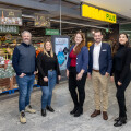Fünf Personen stehen lächelnd vor dem Eingang eines Supermarkts mit der Aufschrift "BILLA PLUS." Sie sind casual bis geschäftlich gekleidet. Im Hintergrund sind Einkaufswagen und Produkte zu sehen.