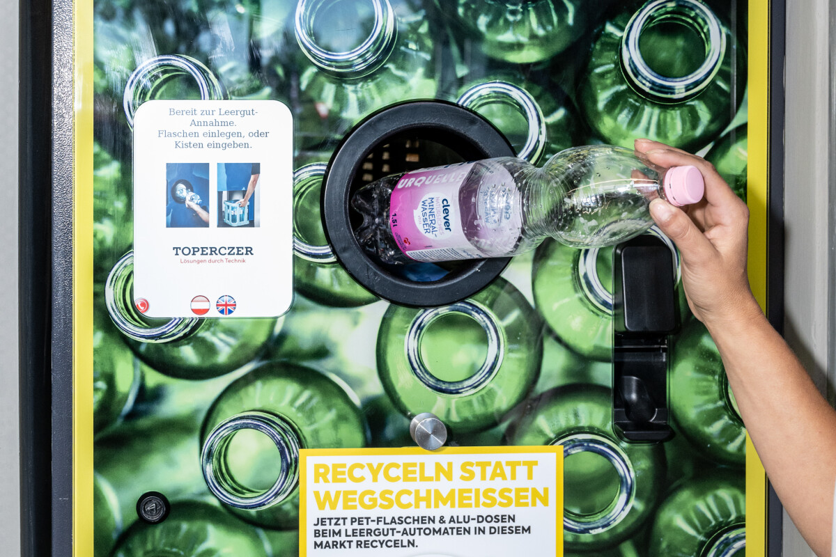 Eine Person steckt eine Plastikflasche in einen Rückgabeautomaten für Leergut. Der Automat ist mit Bildern von Flaschen dekoriert und trägt Aufschriften wie "Leergut Rückgabe" und "Recyceln statt Wegschmeißen".
