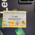 Schild an einem Pfandautomaten mit der Aufschrift: "Dein Pfand fürs Land". Text beschreibt, dass Spenden direkt in regionale Naturschutzprojekte in Österreich fließen. Unten befinden sich Logos mit Blumen und einem Schmetterling.