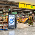 Der Eingang zu einem BILLA PLUS Supermarkt mit sichtbaren Obst- und Gemüseständen. Ein Werbeplakat steht am Eingang. Ein Weihnachtsbaum steht auf der rechten Seite.