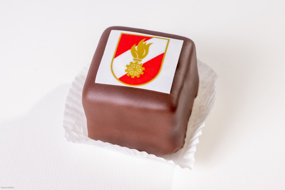 Ein quadratisches Gebäck mit Schokoladenüberzug und einem Wappen mit rotem, weißem Hintergrund und goldenen Symbolen auf einer essbaren Papierdekoration.