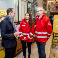 Drei Personen stehen in einem Geschäftsumfeld. Zwei tragen rote Jacken mit einem Rotkreuz-Logo. Sie unterhalten sich mit einem Mann in einem Anzug. Im Hintergrund gibt es Informationsstände und Spendenaufrufe.