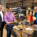 Vier Personen stehen an einer Supermarktkasse. Zwei Männer stehen hinter einem Einkaufswagen, der mit Einkaufstaschen gefüllt ist. Eine Frau und ein weiterer Mann stehen hinter der Kasse. Alle lächeln.
