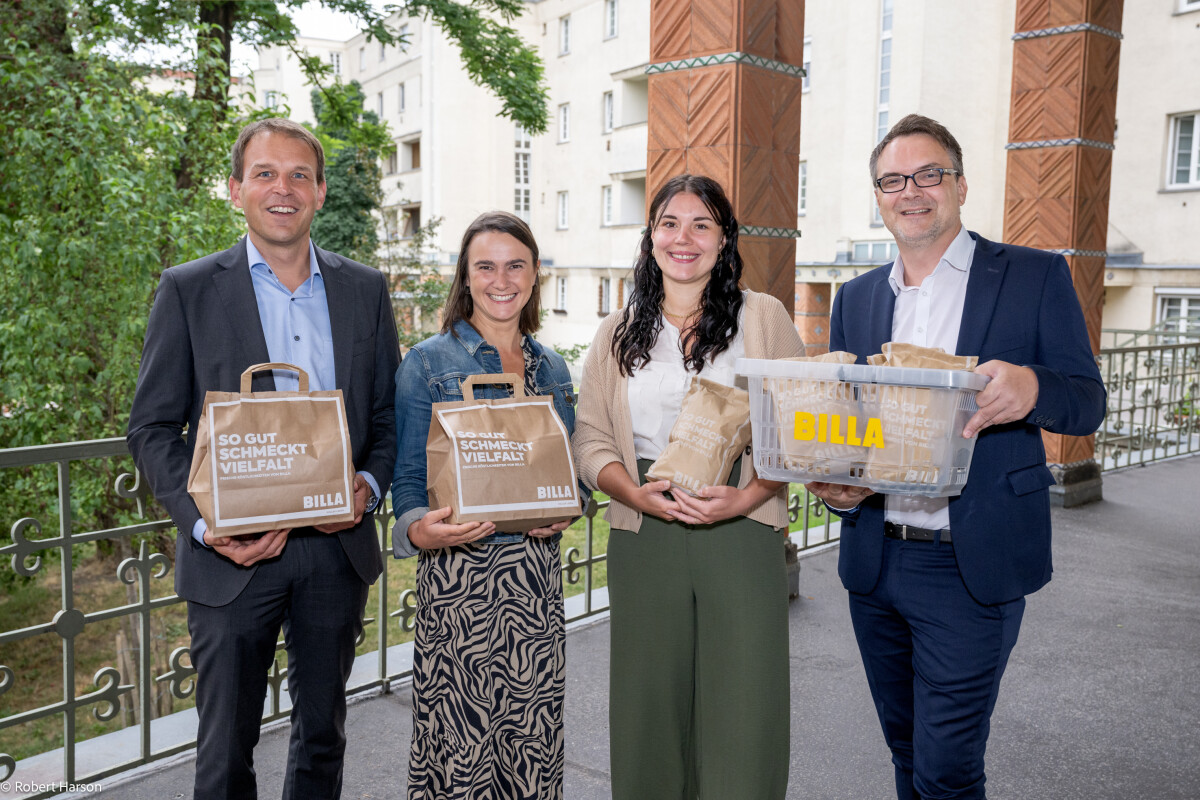 Vier Personen stehen auf einem Balkon und halten Einkaufstaschen von Billa in den Händen. Sie lächeln in die Kamera und tragen Geschäftskleidung. Im Hintergrund sind Bäume und ein Gebäude zu sehen.