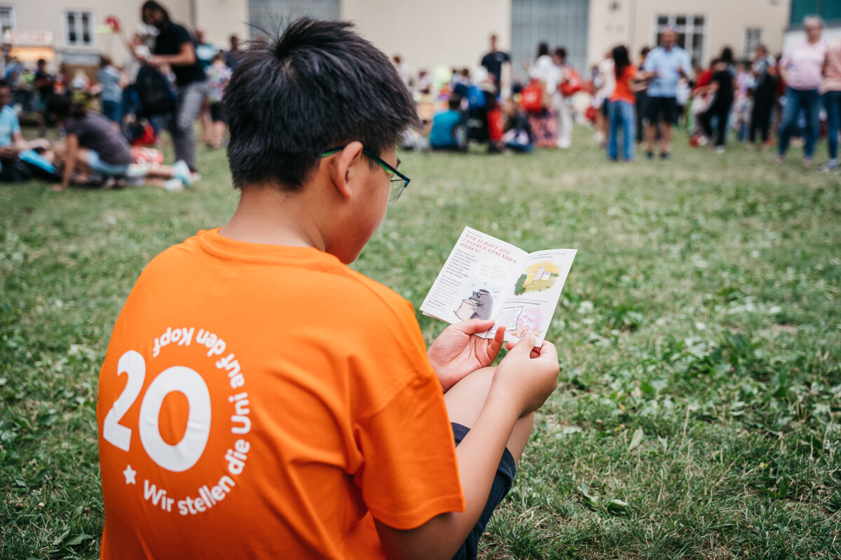 Ein Junge sitzt auf einer Wiese und liest ein Buch. Er trägt ein orangefarbenes T-Shirt mit dem weißen Aufdruck "20". Im Hintergrund sind mehrere Personen und Bäume zu sehen.