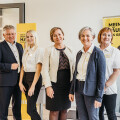 Fünf Personen lächeln in Büro- oder Veranstaltungskleidung. Im Hintergrund befinden sich Werbeplakate in Gelb mit Text über Ausbildung und Jobs. Ein Mann trägt einen Anzug, die Frauen tragen Blusen und Jacken.