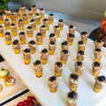 Eine Vielzahl kleiner Dessertgläser, gefüllt mit einer cremigen Masse und Beeren, auf einem Tisch arrangiert. Im Hintergrund ist ein Turm aus bunten Macarons zu sehen.