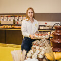 Eine Frau in weißer Bluse hält ein Tablett mit Desserts in einem Buffetbereich. Vor ihr stehen Gläser mit Nachtisch und ein Schokoladenbrunnen. Im Hintergrund sind verschiedene Speisen aufgereiht.