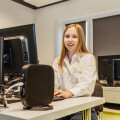 Eine lächelnde Frau sitzt an einem Schreibtisch mit Computern in einem Büro oder Klassenzimmer. Sie trägt ein weißes Hemd und hat einen Namensaufkleber.