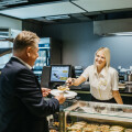 Ein Mann im Anzug erhält eine Mahlzeit von einer lächelnden Angestellten hinter einer Theke in einem modernen Café oder Bistro. Im Hintergrund sind Sandwiches und Gebäck in einer Vitrine ausgestellt.