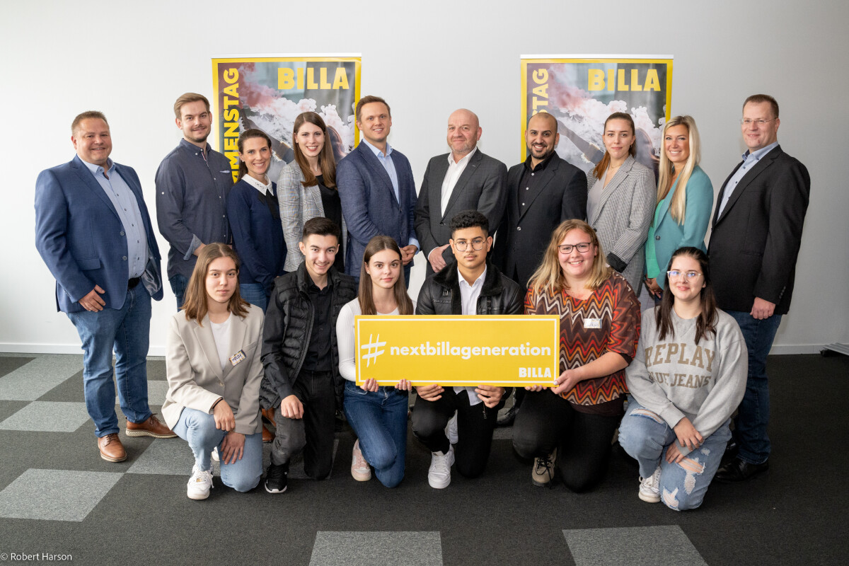 Eine Gruppe von Menschen steht zusammen für ein Foto. Im Vordergrund hält eine Frau ein gelbes Schild mit der Aufschrift "#nextbillageneration BILLA". Im Hintergrund sind zwei Plakate mit dem Wort "BILLA" sichtbar. Alle Personen lächeln.