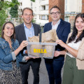 Vier Personen stehen auf einem Balkon und halten zusammen eine Plastikbox mit der Aufschrift "BILLA", die mit Papiertüten gefüllt ist. Sie lächeln in die Kamera. Im Hintergrund sind Gebäude und Bäume sichtbar.
