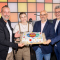 Eine Gruppe von vier Personen hält gemeinsam eine Jubiläumstorte mit der Aufschrift "70". Alle lächeln, während sie die Torte präsentieren. Im Hintergrund ist eine bunte Wand zu sehen.