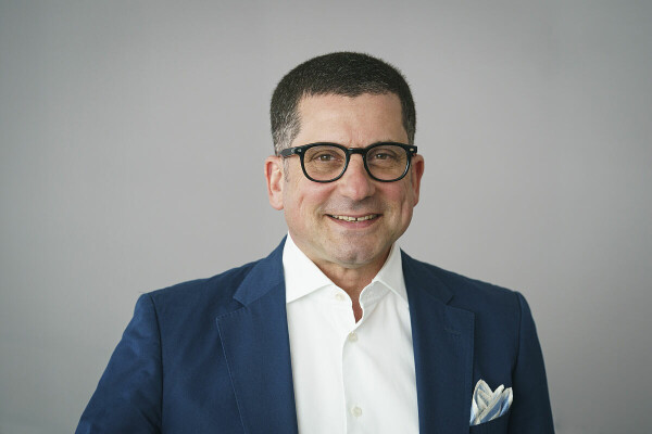 Ein lächelnder Mann mit Brille, trägt einen blauen Anzug und ein weißes Hemd vor einem grauen Hintergrund.