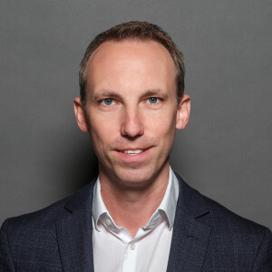 Ein Mann in einem dunklen Anzug mit weißem Hemd lächelt leicht vor einem grauen Hintergrund.