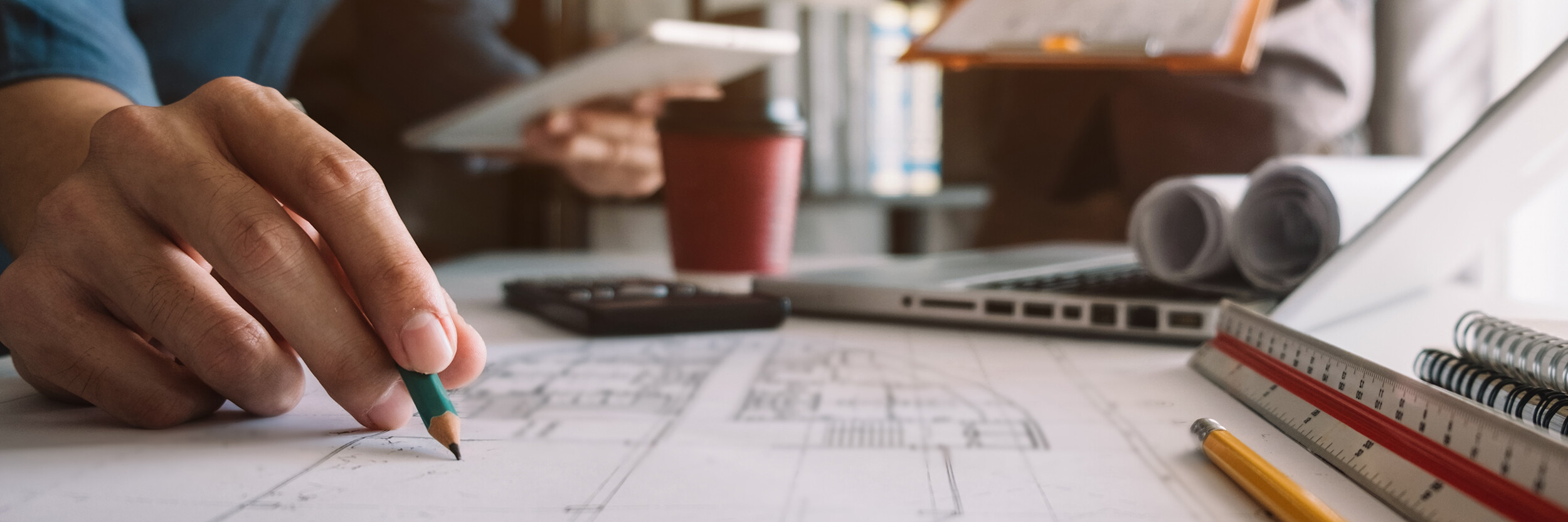 Nahaufnahme einer Hand, die auf einem Architektenplan schreibt. Im Hintergrund stehen ein Laptop, eine Kaffeetasse und zwei Personen, wovon eine ein Tablet hält. Es sind ein Taschenrechner, ein Maßstab und verschiedene Papierrollen zu sehen.