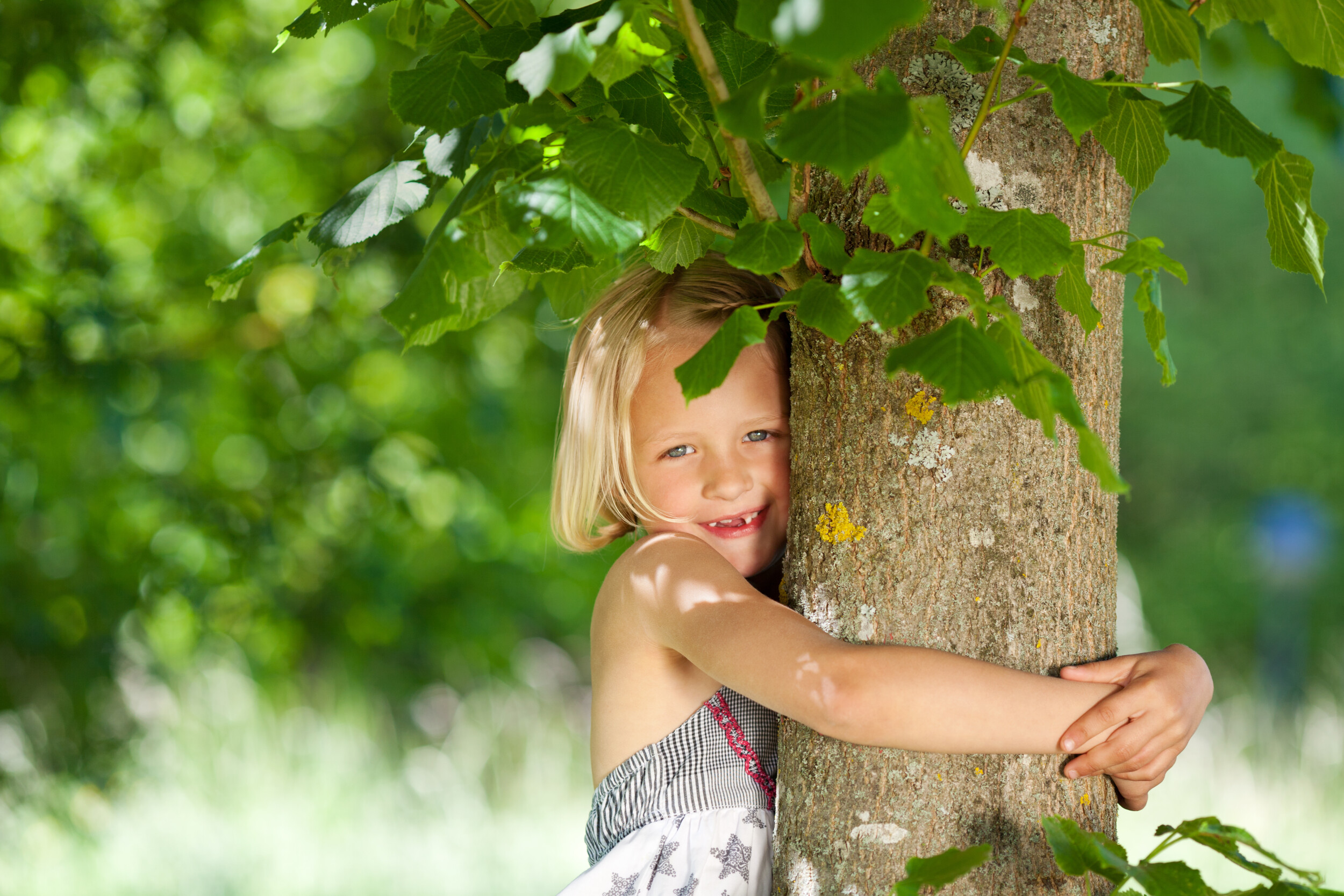 Ein blondes Mädchen umarmt einen Baum und lächelt, umgeben von grünem Laub und hellem Sonnenlicht.
