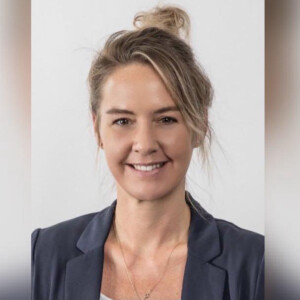 Eine lächelnde Person mit blondem Haar und Dutt trägt einen dunklen Blazer vor einem hellen Hintergrund.