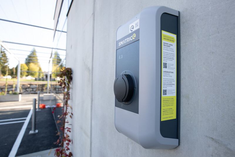 Ladestation für Elektrofahrzeuge an einer Betonwand. Textinformationen auf der Station. Im Hintergrund sind leere Parkplätze und Bäume zu sehen.