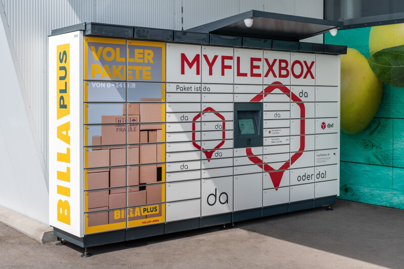 Paketstation mit der Aufschrift "MYFLEXBOX" und "BILLA PLUS". Enthält ein digitales Display und mehrere Schließfächer. Im Hintergrund ist ein großer Apfel sichtbar.