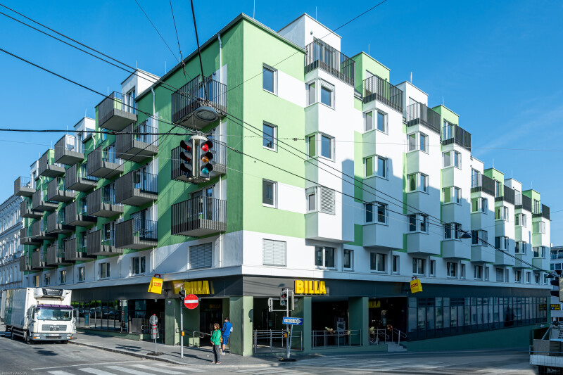Mehrstöckiges Wohn- und Geschäftsgebäude mit grünen und weißen Fassaden. Unten befindet sich ein Supermarkt der Marke Billa. Mehrere Balkone ragen hervor. Ein Lastwagen steht vor einem Fenster. Ampeln und Straßenkreuzung im Vordergrund. Der Himmel ist klar und blau.