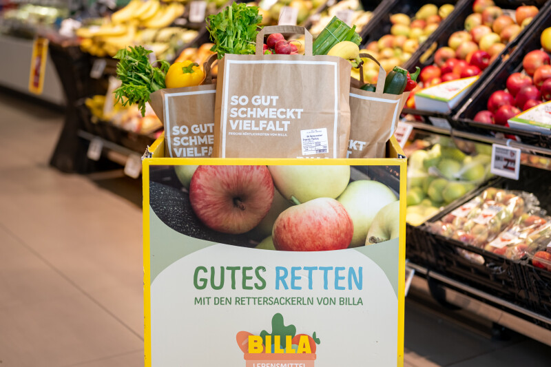 Ein Einkaufswagen voller Gemüse und Obst sowie Papiertüten mit der Aufschrift "So gut schmeckt Vielfalt". Im Hintergrund sind Obst- und Gemüsestände in einem Supermarkt zu sehen. Auf dem Einkaufswagen steht "Gutes retten" mit dem Logo von Billa.