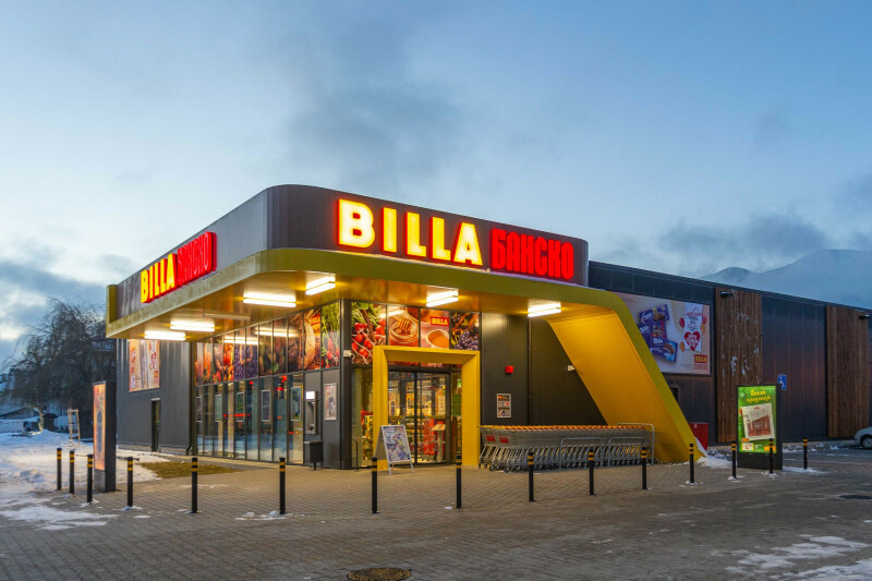 Ein BILLA-Supermarkt mit leuchtendem roten und gelbem Schild in einem modernen Gebäude. Es ist abends, der Himmel ist leicht bewölkt und Schnee liegt auf dem Boden. Fahrradbügel und Einkaufswagen befinden sich vor dem Eingang.