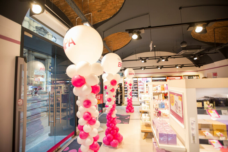Innenraum eines Geschäfts mit hoher Decke und modernen Lichtelementen. Weiße und rosa Ballons sind zu Säulen geformt. Regale mit Kosmetik- und Pflegeprodukten sind sichtbar. Ein großes Schaufenster zeigt die Straße draußen.