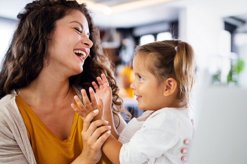 A woman with long curly hair smiling and playing with a young girl indoors. Both appear happy and engaged with each other.