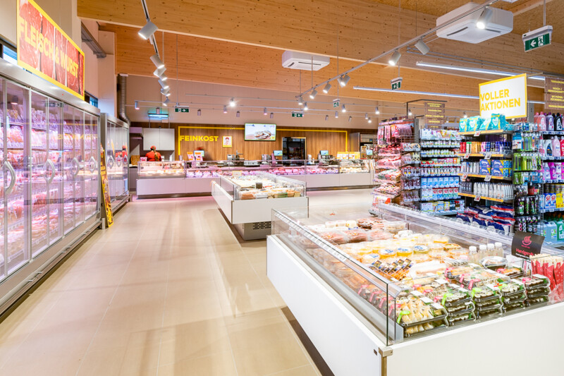 Wide-angle view of a brightly lit supermarket interior, featuring aisles with colorful packaged goods, a refrigerated section to the left, and a display with a variety of foods in the center. The ceiling is wooden with exposed beams, and clear signage is visible overhead.