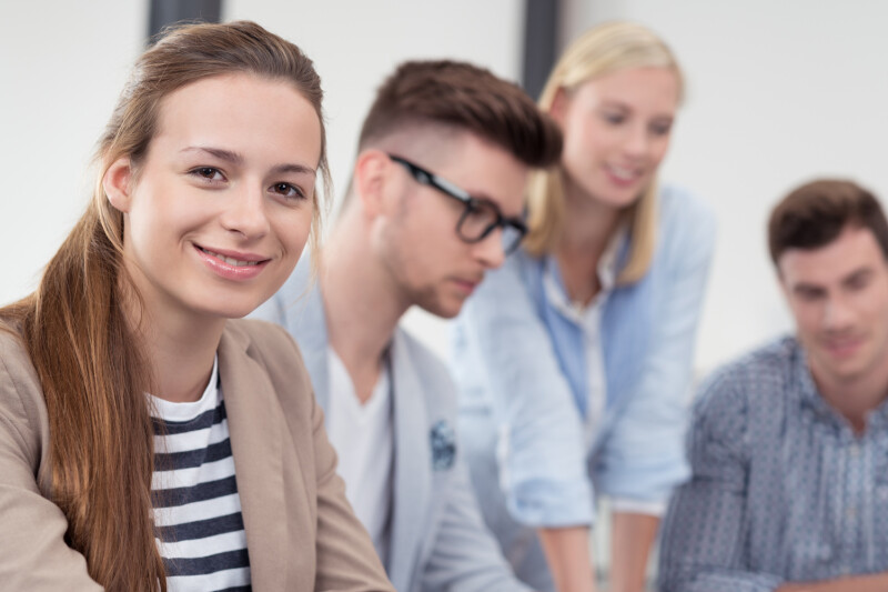 A group of four young adults in an office setting. A woman in the foreground smiles at the camera, wearing a striped shirt and beige blazer. Three colleagues, two men and one woman, are in the background, engaged in a discussion.