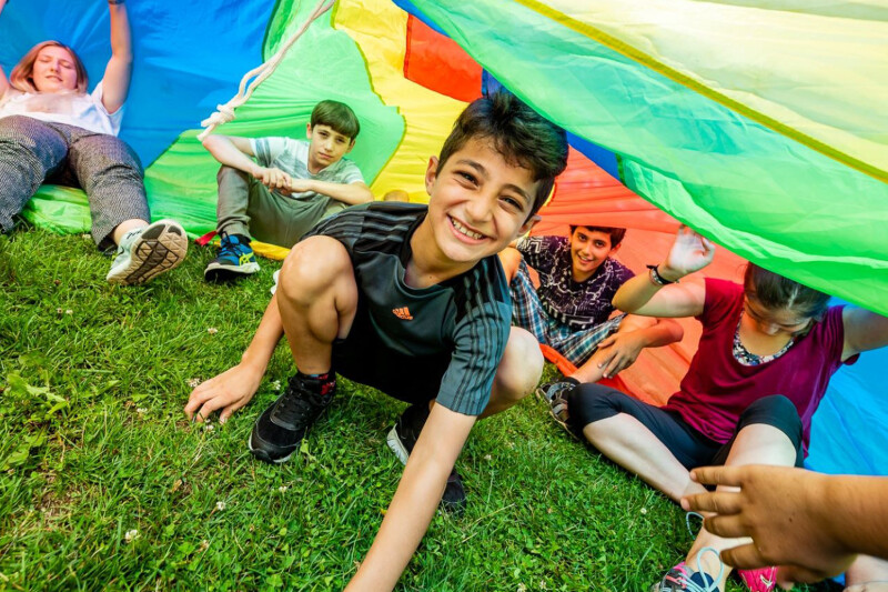 Children playing under a colorful parachute, smiling and crouching on the grass. One child reaches out with their hand visible in the foreground.