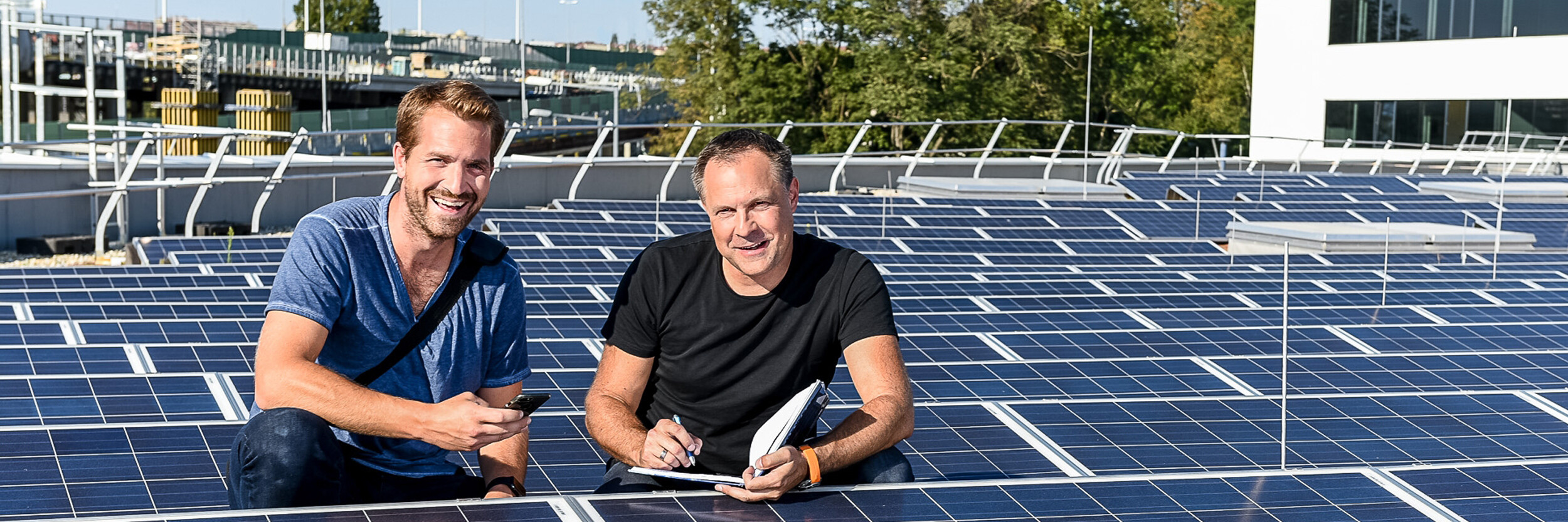 Two men sitting on a rooftop covered with solar panels, smiling and engaging with a smartphone and a notebook. A building and greenery are visible in the background.