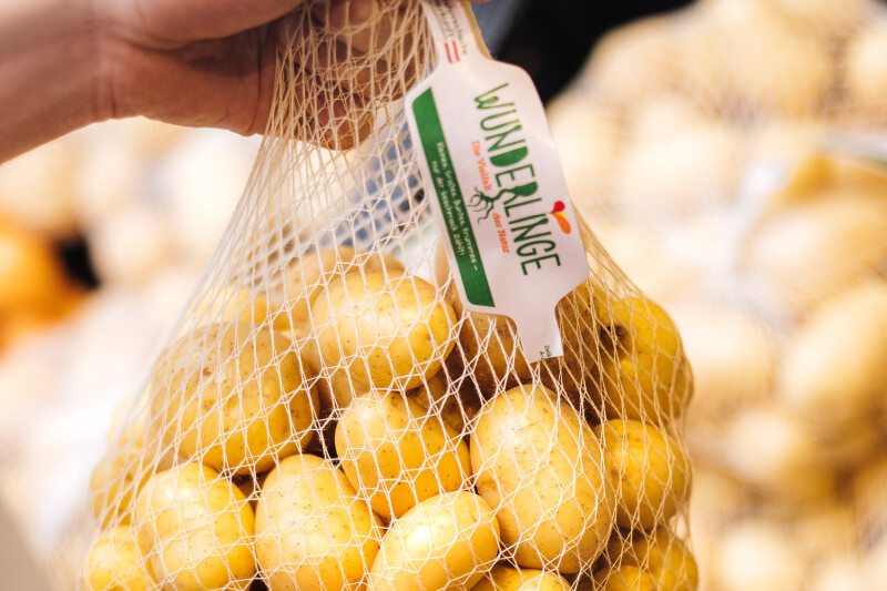 A hand holds a mesh bag filled with yellow potatoes, labeled "Wunderlinge." The background shows more bags of potatoes slightly out of focus.