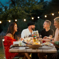 A group of people enjoying an outdoor dinner party at dusk. The table is decorated with food and drink, and string lights hang above, creating a warm atmosphere. The people are smiling and engaged in conversation.