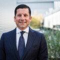 A man in a dark pinstripe suit and tie stands smiling outdoors, with a blurred background of plants and modern building architecture.