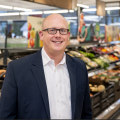 A person in a suit and glasses stands smiling in a brightly lit grocery store aisle, with various fruits and vegetables displayed behind them.