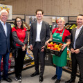 Five people stand in a grocery store produce section. The person in the center holds a box of bell peppers. They are surrounded by displays of fruits and vegetables. All are dressed in business or store attire, smiling at the camera.